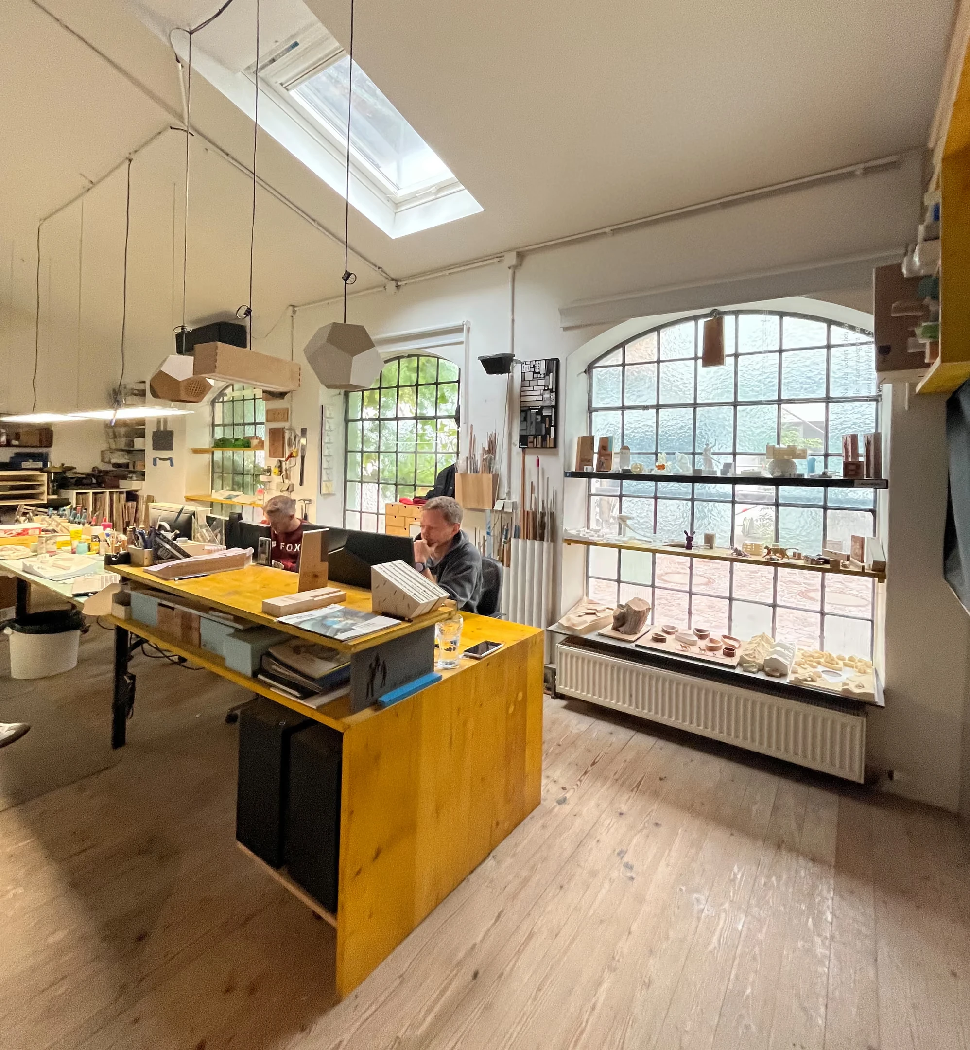 Blick ins Atelier mit Rundbogenfenstern, Regalen voller Modelle und zwei Personen am gelben Arbeitstisch.